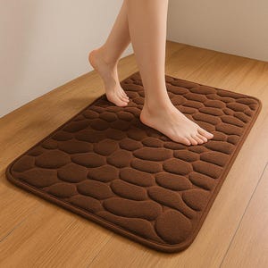 May include: A rectangular brown bath mat with a pebble design. The mat is made of a soft, plush material. A person is stepping onto the mat with bare feet. The mat is on a wooden floor.