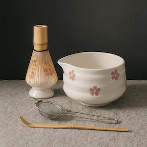 Op de afbeelding: Een matcha-theeset met een witte keramische kom met roze bloemmotieven, een bamboe klopper, een metalen zeef en een bamboe lepel. De set staat op een grijze ondergrond.