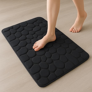 May include: A black rectangular bath mat with a pebble design. The mat is made of a soft material and is designed to absorb water. The mat is approximately 24 inches long and 16 inches wide.