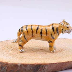 May include: A small tiger figurine with an orange body and black stripes. The tiger stands on a round wooden base. The figurine's paws are silver-coloured. The wooden base is light brown. The figurine is a detailed representation of a tiger.