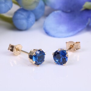 May include: A pair of gold stud earrings with round, deep blue gemstones. The earrings are set against a white background with blue floral accents, showcasing the jewelry's color and design. The studs are a classic jewelry item.