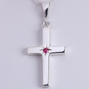 May include: A silver cross pendant featuring a small red gemstone at its centre. The cross is simple and elegant, making it a perfect addition to any jewellery collection.