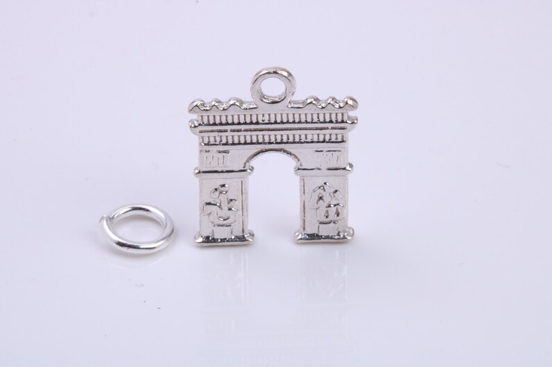 May include: Silver charm in the shape of the Arc de Triomphe, a famous Parisian landmark. The charm has a loop at the top for attaching to a bracelet or necklace.