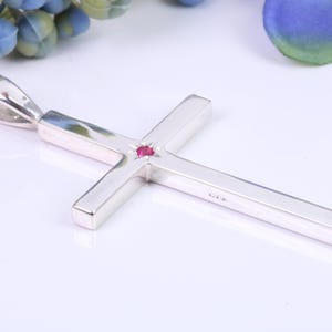 Puede incluir: Un colgante de cruz de plata con una pequeña piedra roja en el centro. El colgante tiene un diseño simple y moderno y es perfecto para un collar.