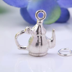 Peut inclure: Un charm argenté en forme de théière avec une boucle pour l'accrocher. La théière a une anse et un bec verseur. Un petit anneau argenté est à côté du charm. Le fond présente des éléments floraux bleus et verts flous.