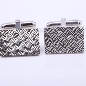 May include: A pair of silver cufflinks with a geometric pattern. The cufflinks are rectangular in shape and have a textured surface.