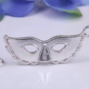Può includere: Un ciondolo a forma di maschera argentata con un piccolo anello accanto. La maschera ha fori per gli occhi e un naso, con un bordo decorativo. Il ciondolo è probabilmente per la creazione di gioielli o l'artigianato.
