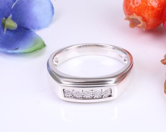 Puede incluir: Un anillo de plata con una cara rectangular engastada con cinco pequeños diamantes de talla cuadrada. El anillo tiene un acabado pulido y un diseño clásico. El fondo incluye pétalos de flores azules y bayas naranjas.
