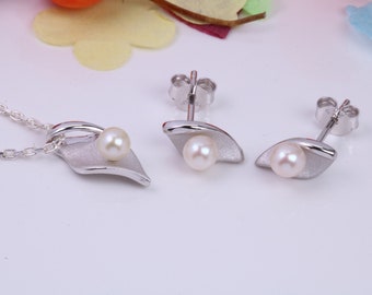 Puede incluir: Un conjunto de collar y pendientes de plata con una perla en cada pieza. El collar tiene una cadena delicada y un colgante con forma de hoja. Los pendientes son pendientes de botón con un diseño en forma de hoja.