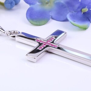 Op de afbeelding: Zilveren kruisvormige hanger met roze edelsteen inleg. Het kruis is hoogglans gepolijst en reflecteert licht, met een kleine lus voor een ketting. De achtergrond heeft blauwe en groene bloemen.