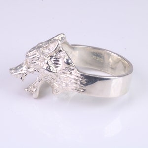 Op de afbeelding: Een zilveren ring met een wolfskopontwerp. De bek van de wolf is open en zijn tanden zijn zichtbaar. De ring is gemaakt van sterling zilver en heeft een gepolijste afwerking.