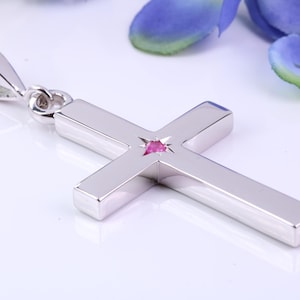 May include: A silver cross pendant with a small, star-shaped pink gemstone at the center. The cross is highly polished and has a loop at the top for a chain. The background is white with a blurred blue and green floral element.