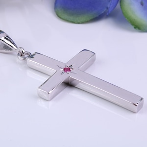 Könnte beinhalten: Silberner Kreuzanhänger mit einem kleinen, roten Edelstein in der Mitte. Das Kreuz ist glatt und poliert, mit einer Schlaufe für eine Kette. Der Hintergrund ist weiß mit einem Hauch von Blau.