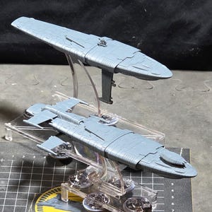 May include: Two gray model spaceships, one above the other, displayed on clear stands. The ships have a detailed design and are set against a dark background. The lower ship is on a black and gray grid.