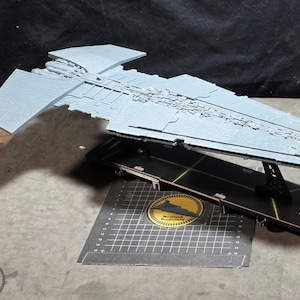 Puede incluir: Maqueta gris claro de una nave estelar tipo destructor, sobre un soporte negro. El modelo presenta detalles intrincados y se encuentra sobre un fondo oscuro. Un emblema circular es visible en la base.