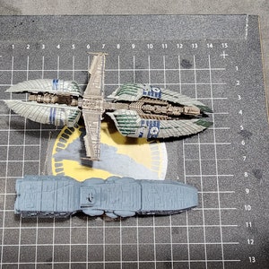 May include: Two model spaceships, one gray and one green and blue, on a gray grid background. The gray spaceship is longer and wider than the green and blue spaceship.
