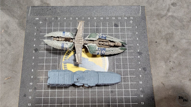 May include: Two model spaceships, one gray and one green and blue, on a gray grid background. The gray spaceship is longer and wider than the green and blue spaceship.