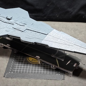 Puede incluir: Modelo gris claro de una nave espacial con intrincados detalles de superficie, apoyado sobre un soporte negro. El modelo tiene una parte delantera larga y puntiaguda y una parte superior ancha y plana. El soporte es negro con detalles amarillos.