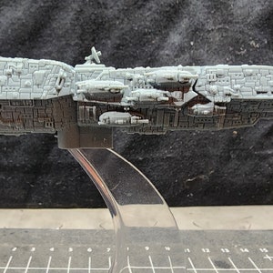 May include: A gray, detailed model of a large spaceship with a long, narrow body and multiple smaller ships attached to the side. The model is on a clear plastic stand.