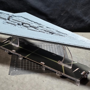 Puede incluir: Maqueta gris de una nave espacial Star Destroyer, apoyada sobre un expositor negro y transparente. La nave espacial tiene detalles de superficie. El expositor tiene una base negra con soportes transparentes. El fondo es oscuro.
