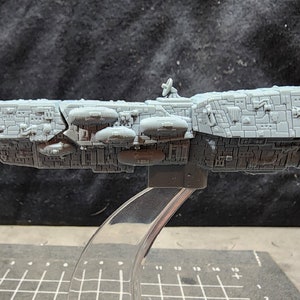 May include: A gray, detailed model of a spaceship with a rounded front and a long, rectangular body. The spaceship has several small, round pods attached to the side and a small, round antenna on top.