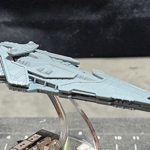 May include: A light gray miniature spaceship model with detailed paneling and a sleek design. The model is mounted on a clear stand, suggesting it's a collectible or display item. The spaceship has a long, pointed front and multiple fins.