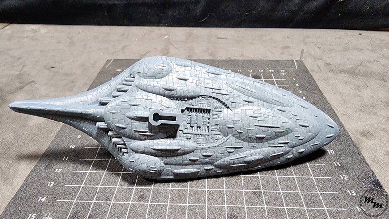 May include: A grey, detailed model of a spaceship with a textured surface and a small hatch on the top. The model is sitting on a grey gridded surface.