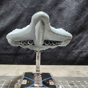 May include: A grey, detailed model of a spaceship with a pointed nose and a row of circular exhaust ports on the underside. The model is mounted on a clear plastic stand.