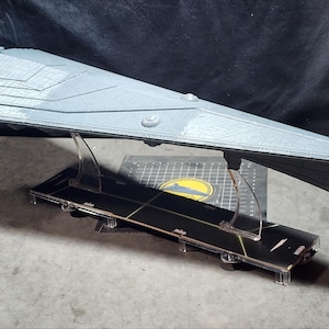 Puede incluir: Maqueta gris de una nave espacial, sostenida por un soporte de acrílico transparente. La nave tiene un cuerpo largo y triangular con paneles detallados y una parte inferior gris oscuro. El modelo se exhibe sobre una superficie gris.