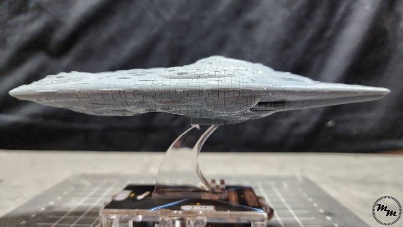May include: A grey, detailed model of a spaceship with a flat, circular base and a rounded top. The model is on a clear plastic stand.