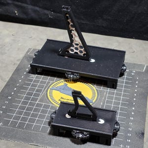 May include: Two black metal display stands. The larger stand has a honeycomb-patterned support. Both stands are on a gray surface with a grid pattern and a yellow and blue emblem.