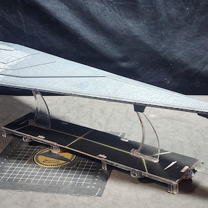 Puede incluir: Modelo gris de una nave espacial Star Destroyer, elevado sobre soportes de acrílico transparente sobre una base negra. La base tiene líneas amarillas y un emblema circular. El modelo es detallado y está sobre un fondo oscuro.