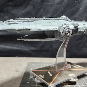 May include: A grey, detailed model of a spaceship on a clear plastic stand. The spaceship has a long, thin body with a pointed nose and a large, round engine at the back. The model is on a black and white chequered surface.