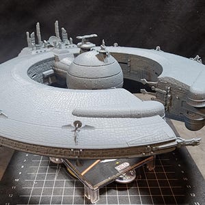 May include: A gray, detailed model of a spaceship with a circular base and a central sphere. The model is made of plastic and has a textured surface.