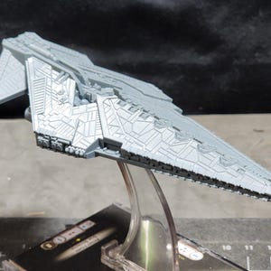 Puede incluir: Maqueta gris de una nave espacial, con detalles intrincados y un diseño triangular. El modelo está montado sobre un soporte transparente, con una base negra visible debajo. La superficie de la nave está cubierta de patrones geométricos.