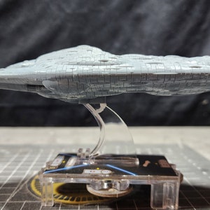 May include: A grey, detailed model of a spaceship, resembling a large, flat, irregularly shaped rock, is displayed on a clear plastic stand. The stand is on a black and grey grid surface.