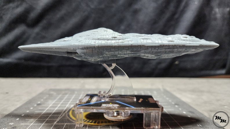 May include: A grey, detailed model of a spaceship, resembling a large, flat, irregularly shaped rock, is displayed on a clear plastic stand. The stand is on a black and grey grid surface.