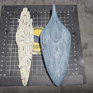 May include: Two model spaceships, one white and one grey, are displayed on a black gridded surface. The white spaceship is long and thin, while the grey spaceship is shorter and wider. Both spaceships have a detailed, textured surface.