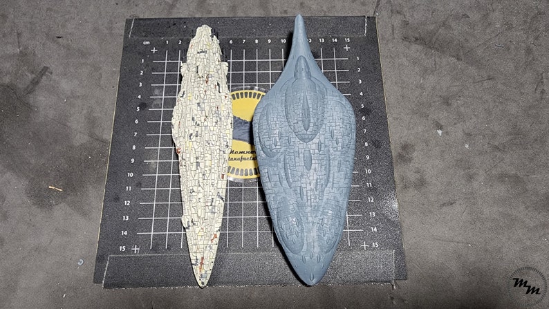 May include: Two model spaceships, one white and one grey, are displayed on a black gridded surface. The white spaceship is long and thin, while the grey spaceship is shorter and wider. Both spaceships have a detailed, textured surface.