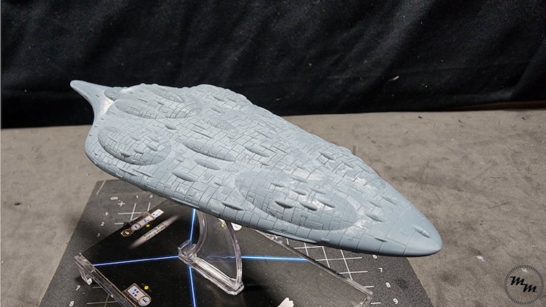 May include: A grey, detailed model of a spaceship with a textured surface. The model is on a clear display stand.