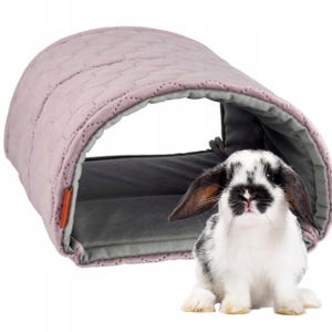 Op de afbeelding: Een roze, gewatteerd tunnelbed voor kleine huisdieren, zoals konijnen, met een grijs, zacht, gevoerd interieur. Een zwart-wit konijn zit voor het bed.