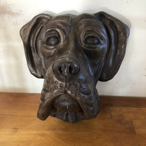 May include: A brown ceramic dog head wall sculpture. The dog has a short snout and large, floppy ears. The sculpture is detailed with realistic features, including wrinkles around the eyes and nose.