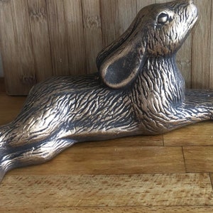 Op de afbeelding: Bronzen konijnenbeeldje in een liggende houding, met het hoofd omhoog en de oren omhoog. De sculptuur heeft een gedetailleerde vachttextuur en staat tegen een houten achtergrond. Het konijn is ongeveer 25 cm lang.