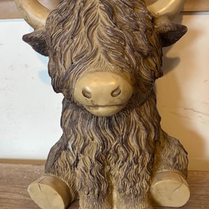 May include: A detailed figurine of a Highland cow, with a shaggy brown coat and tan horns. The cow is in a seated position, with its legs and face clearly visible. The figurine is made of a solid material and has a matte finish.