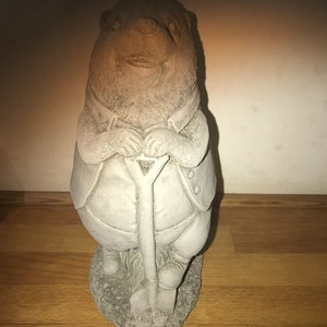 May include: A gray concrete garden statue of a bear wearing a vest and holding a shovel. The bear is standing on a base of concrete.
