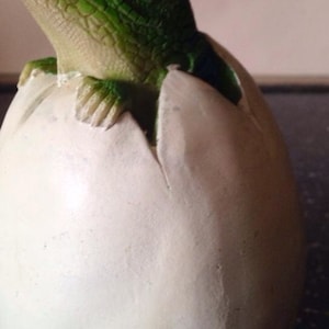 May include: A green dinosaur figurine hatching from a white egg. The dinosaur has a textured green body and a large, open mouth.