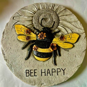 May include: A round, grey stone garden stepping stone with a 3D bee design. The bee is black and yellow with brown and yellow flowers. The stone has the words "Bee Happy" written on it.