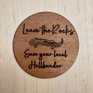 May include: A round wooden coaster with the text "Leave the Rocks Save your local Hellbender" and a black silhouette of a salamander.