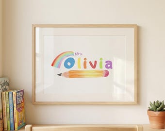 May include: A framed wall print featuring the text Mrs. Olivia in colorful letters, accompanied by a watercolor-style rainbow and a yellow pencil graphic. The artwork is displayed in a light-toned wooden frame on a plain wall.