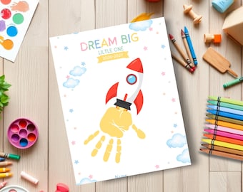 May include: A white rectangular craft sheet featuring a cartoon rocket ship illustration with a yellow handprint as the exhaust flame. The text reads DREAM BIG LITTLE ONE 2026-2027. The design includes light blue clouds and small pink and blue stars.